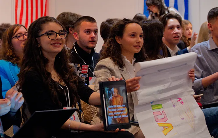 A group of seated delegates smile and hold up materials during the debate portion of the general assembly.