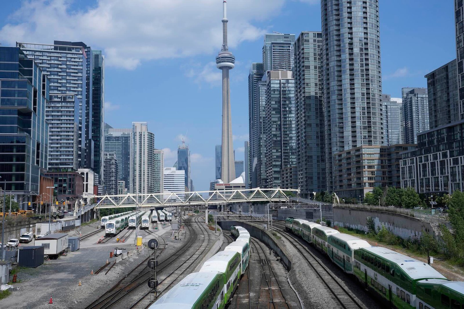 View of multiple train tracks and green trains in downtown Toronto, with high-rise buildings and the CN Tower in the background under a partly cloudy sky.