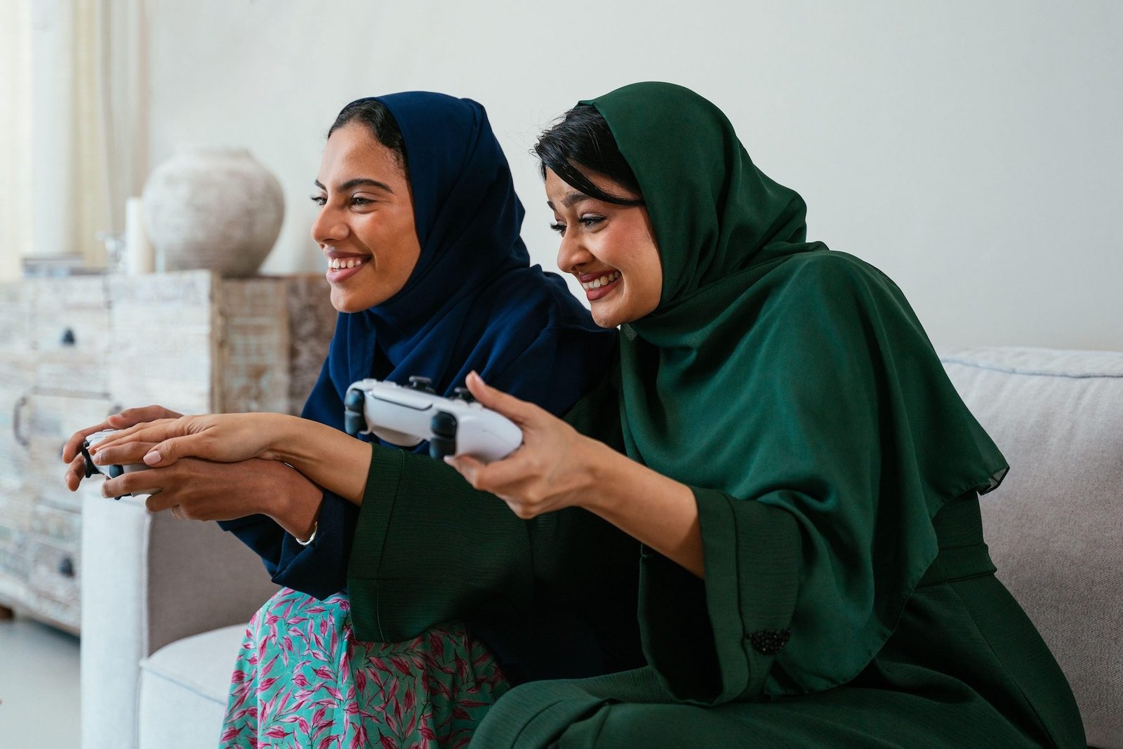 3C3MG74 Two emirati female friends wearing abaya are sitting on a sofa in their living room, enjoying their leisure time by playing video games together and h