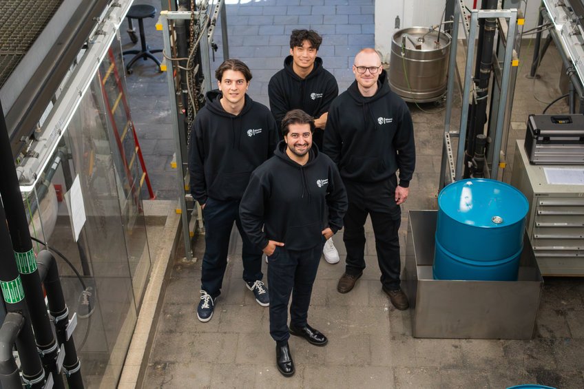 Four people are standing in an industrial hall wearing black hoodies with logos. Next to them is a blue barrel and technical equipment.
