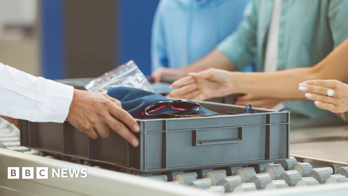 A passenger is placing items into a grey tray at airport security as another pair of hands, presumably a security staff member, holds onto the tray on the other side.