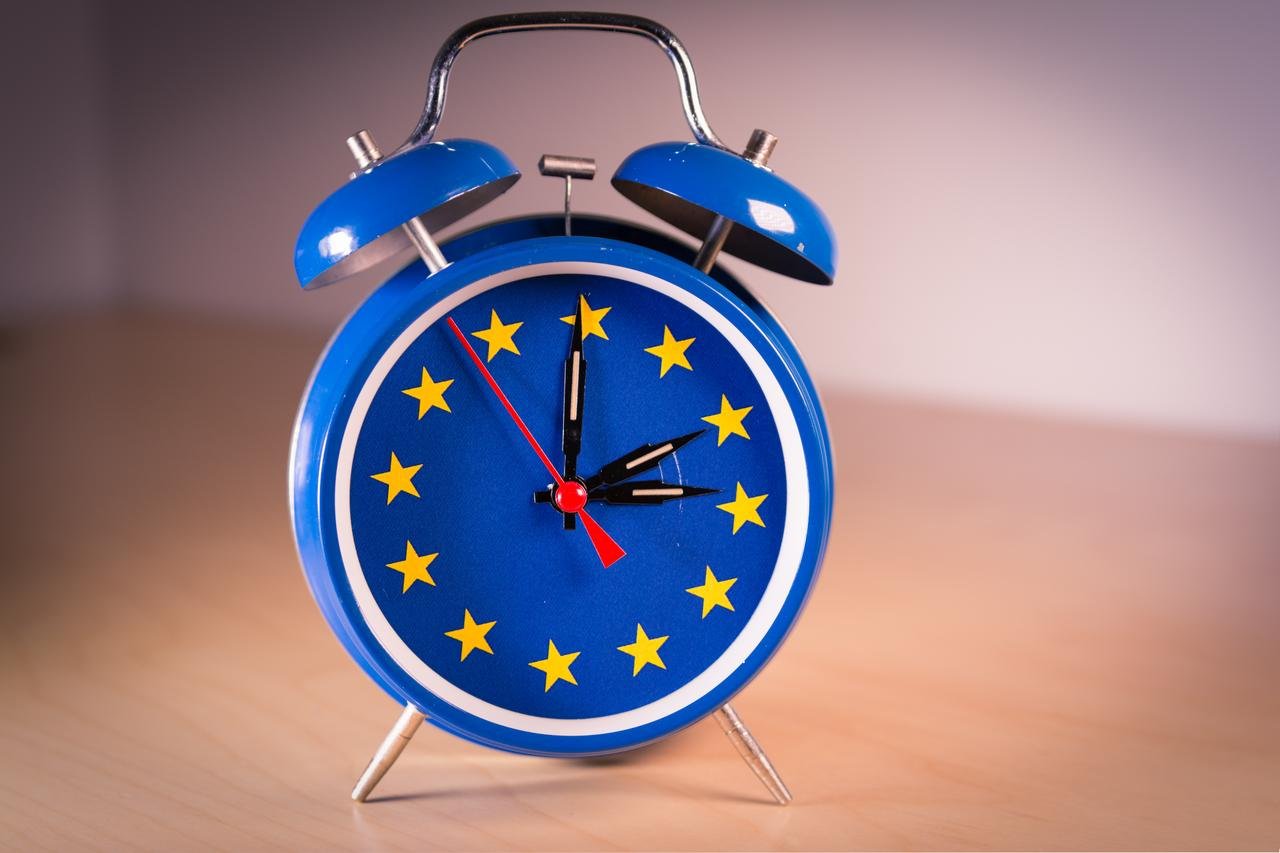 A vintage-style alarm clock designed in the colors of the EU flag, with its hour and minute hands moving forward or backward by one hour to symbolize the time change in Europe. (Adobe Stock Photo)