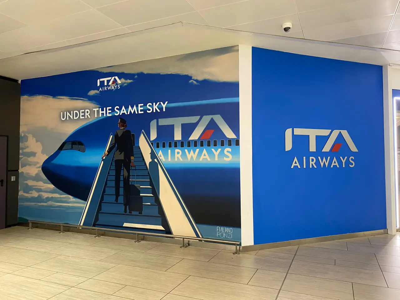 ITA Airways wall mural in an airport terminal showing a pilot boarding a blue aircraft with the slogan “Under the Same Sky.”
