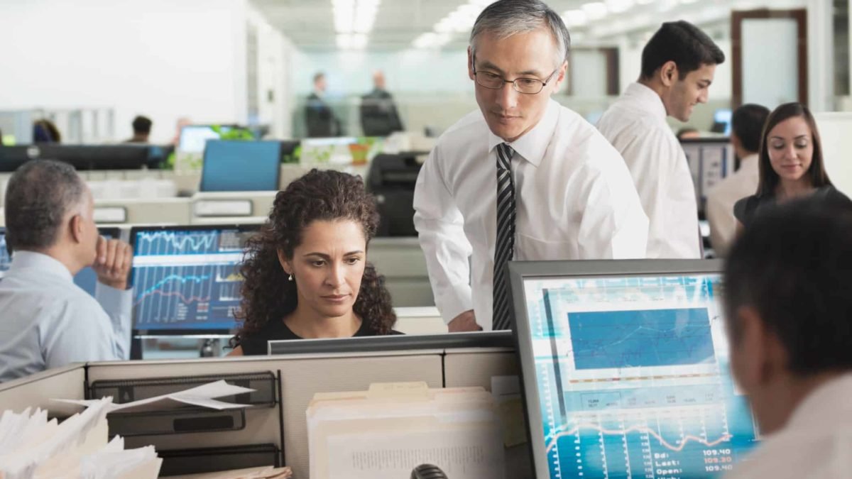 A group of market analysts sit and stand around their computers in an open-plan office environment.