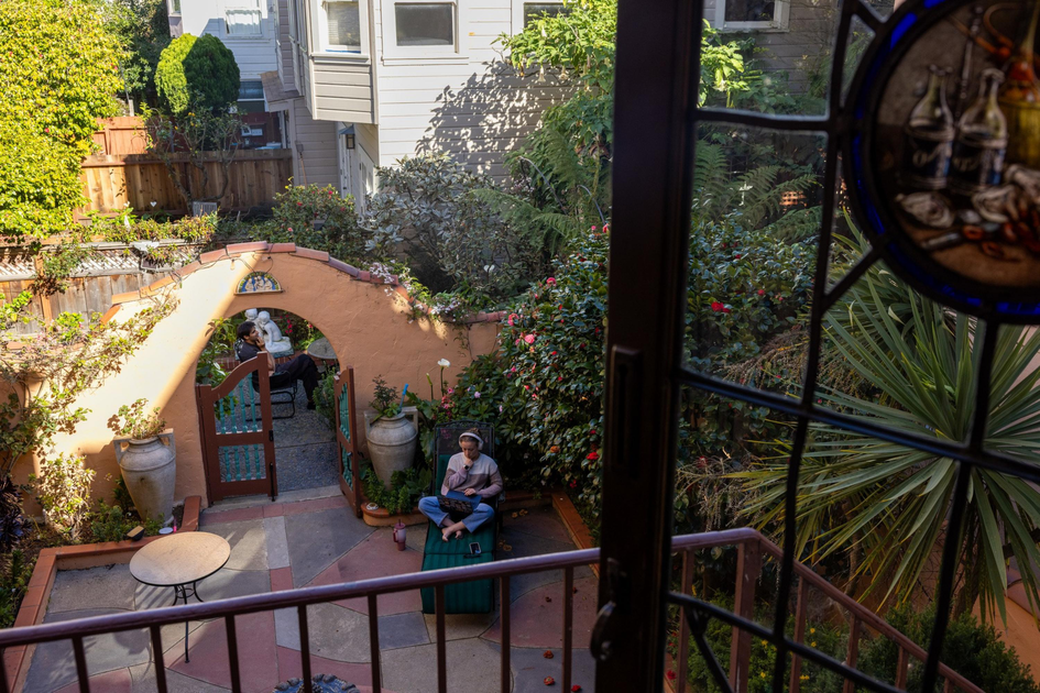A person sits cross-legged on a green bench in a sunlit garden patio, surrounded by plants, flowers, large pots, an archway, and another person beyond it.