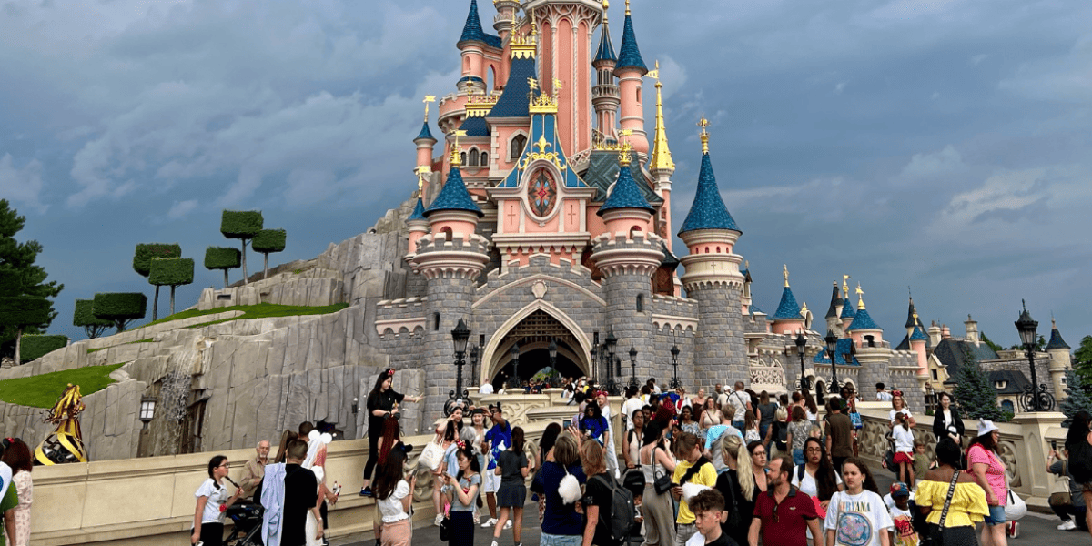 A bustling scene of visitors at Disneyland Paris in front of the iconic Sleeping Beauty Castle, with its pink and blue spires under a cloudy sky.