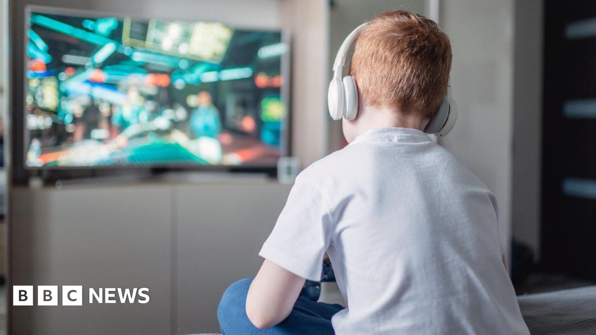 A boy with ginger hair is sitting down with his back to the camera, playing a video game on a large tv in the background. He has white headphones on and is wearing a white t-shirt and blue jeans.