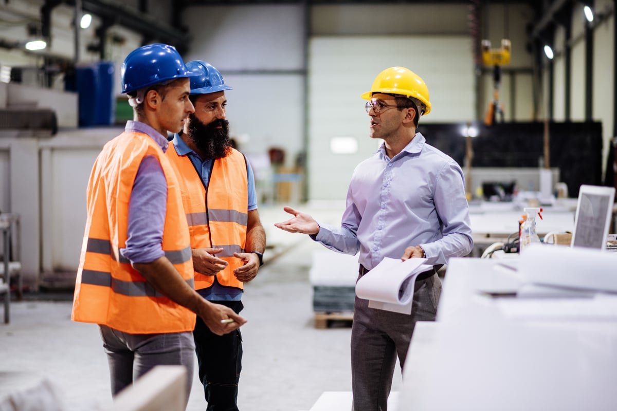 The image shows engineers wearing hard hats talking inside a factory hall.