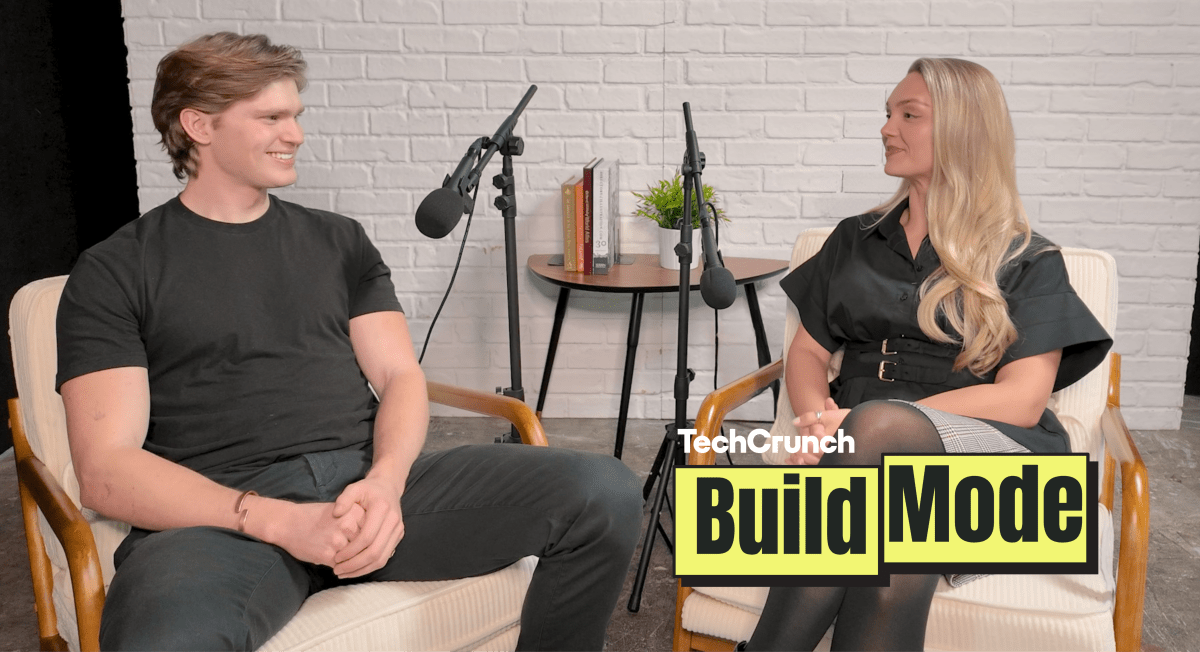 Artisan CEO, Jaspar Carmichael-Jack and Build Mode host Isabelle Johannessen sit facing each other in a podcast studio with microphones, mid-conversation, in front of a white brick wall; a small table with books and a plant sits between them, and a “TechCrunch Build Mode” logo overlay appears in the lower right corner.