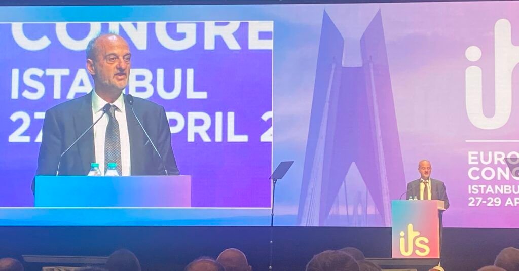 A speaker addresses delegates from a podium at the ITS European Congress in Istanbul, with the congress branding and city logo visible on a large screen behind him, and a second speaker visible at a podium in the background to the right