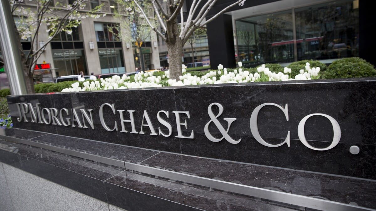 A large black stone sign with silver letters reads JPMorgan Chase & Co. in front of a city office building with white flowers and greenery in the background.