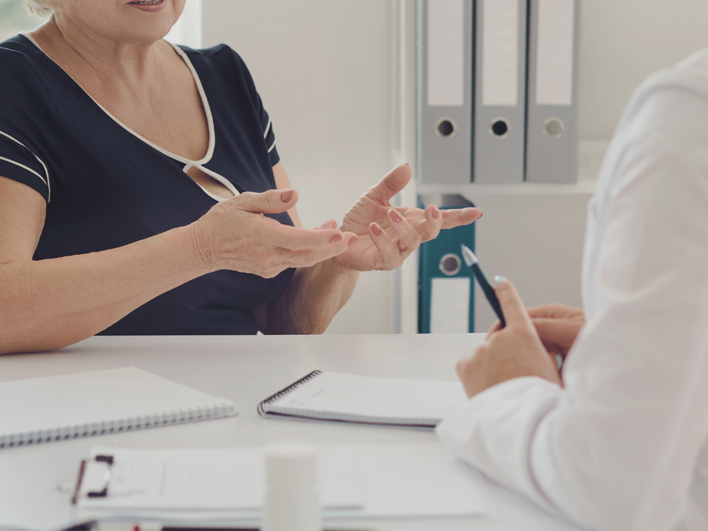 An older woman sitting across from a doctor at a desk during a medical consultation. The woman is gesturing with her hands while speaking. The doctor, holding a pen, listens attentively. Notebooks, documents, and a prescription bottle are on the desk, with shelves of binders in the background.