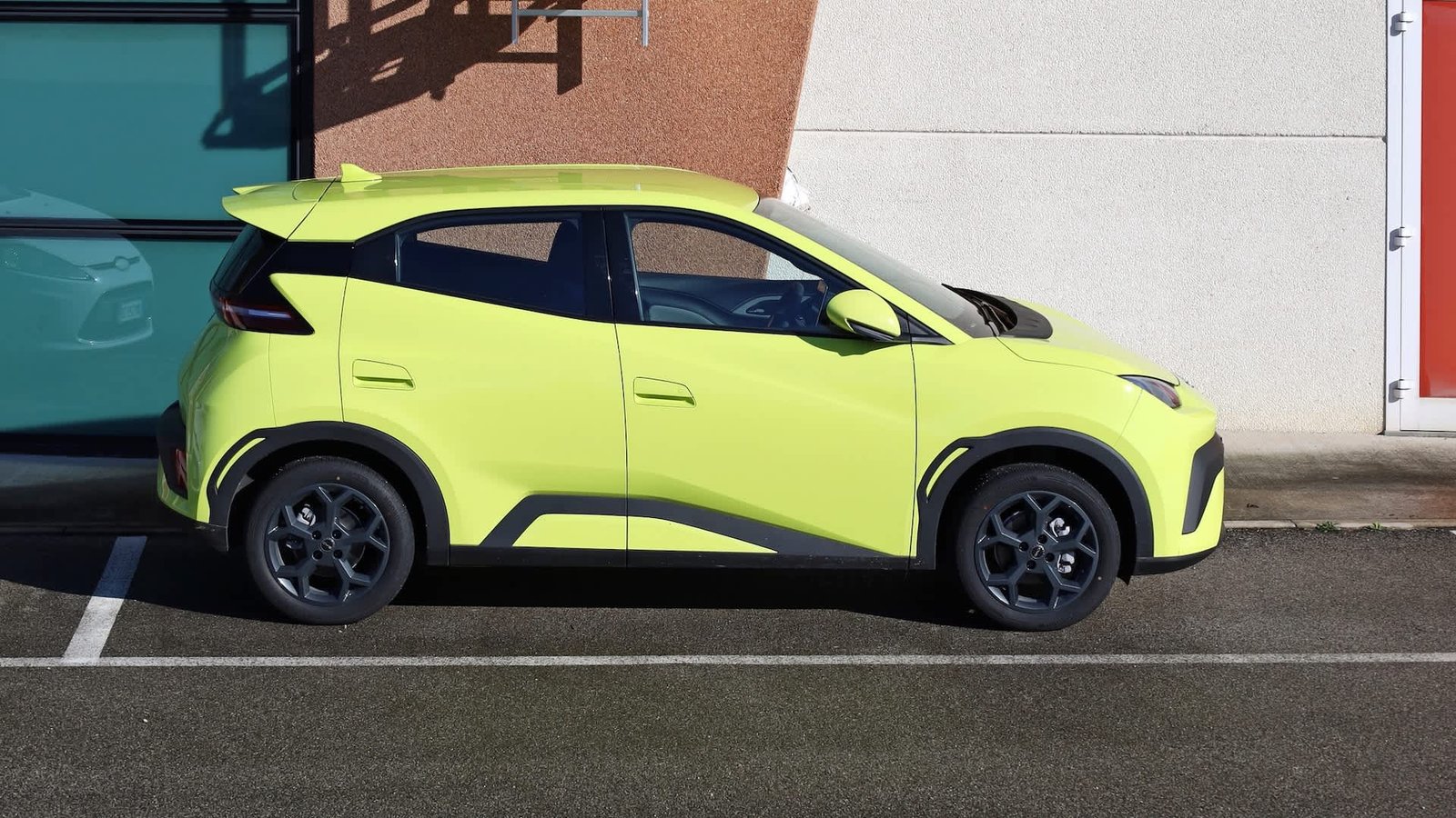 A bright yellow compact car parked beside a textured wall and glass windows in an urban setting.