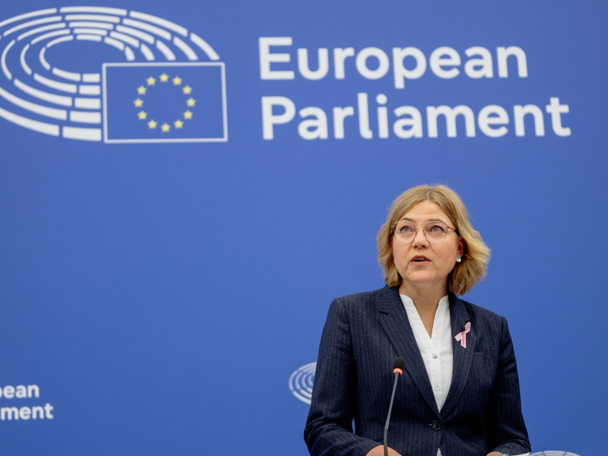 Henna Virkkunen wears a dark blazer and white shirt and stands at a lectern in front of a blue wall with European Parliament written on it.