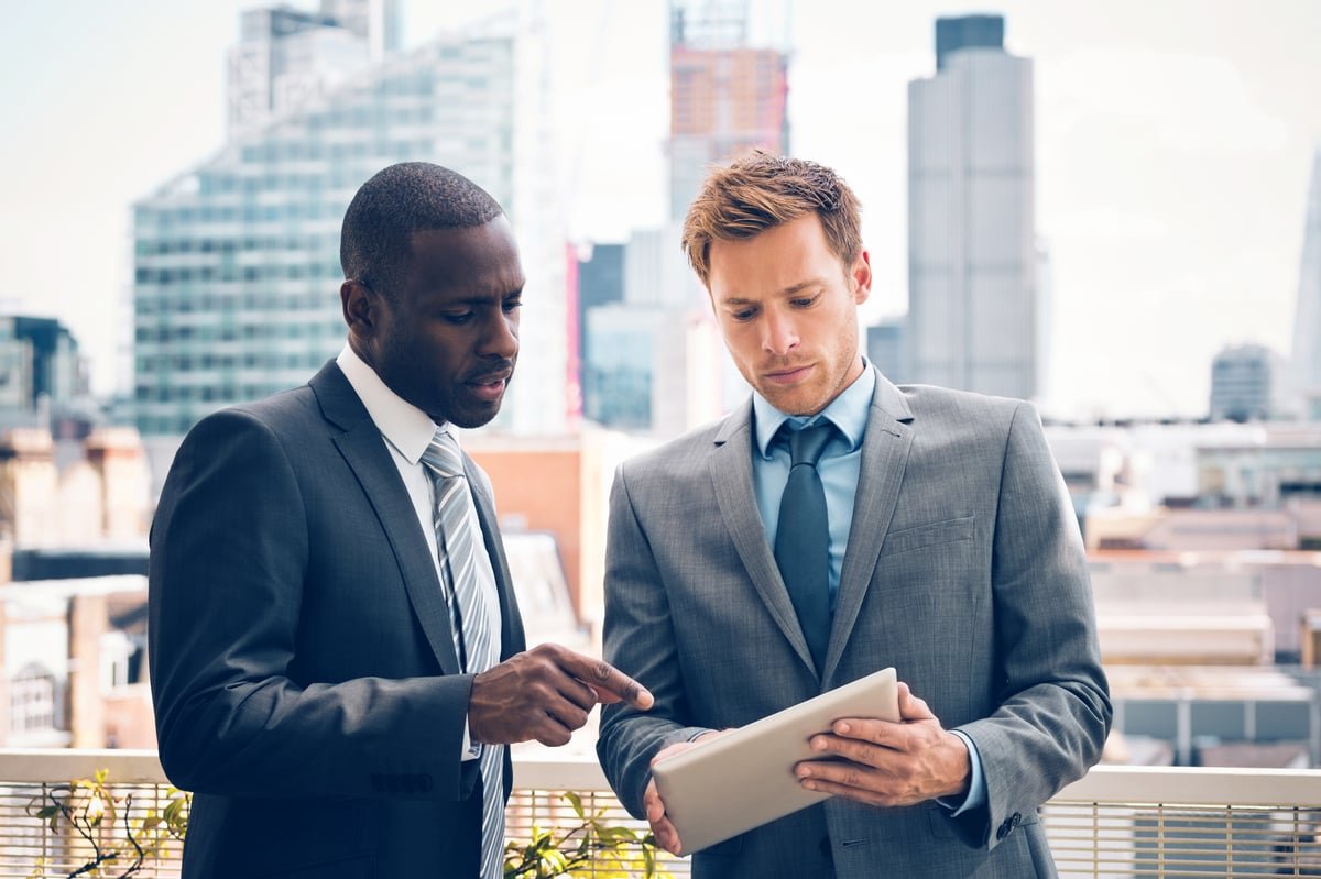 Two investors stand on a roof deck while one shows data on a tablet computer to the other.