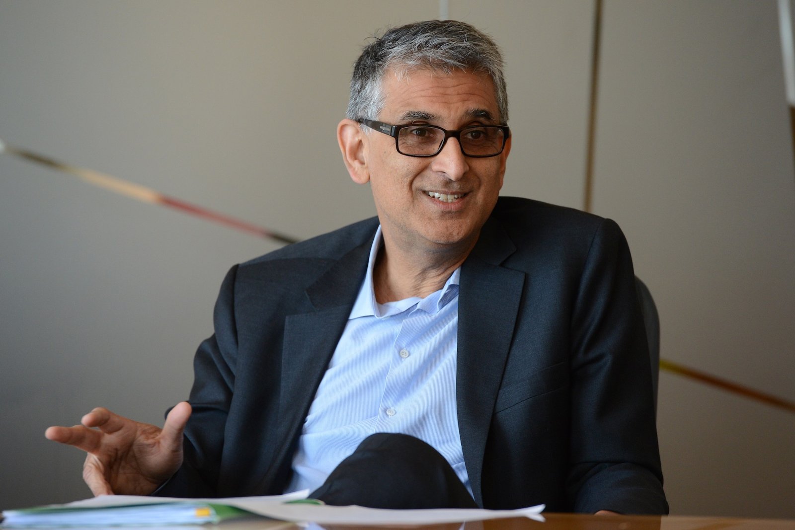 A portrait-style shot of Nadir Mohamed wearing a dark suit, with a blue shirt and no tie. He's seated at a desk and has a slight smile on his face.