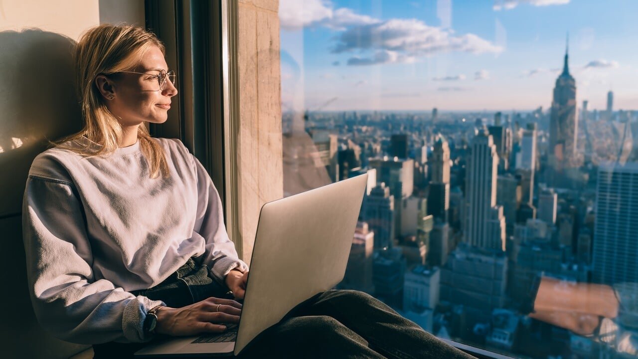 Caucasian female digital nomad sitting with laptop computer on window sill of modern skyscraper building coworking