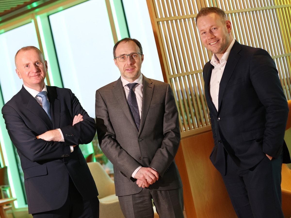 Three men in suits pose for the camera. They are standing in an office setting with a window behind them.