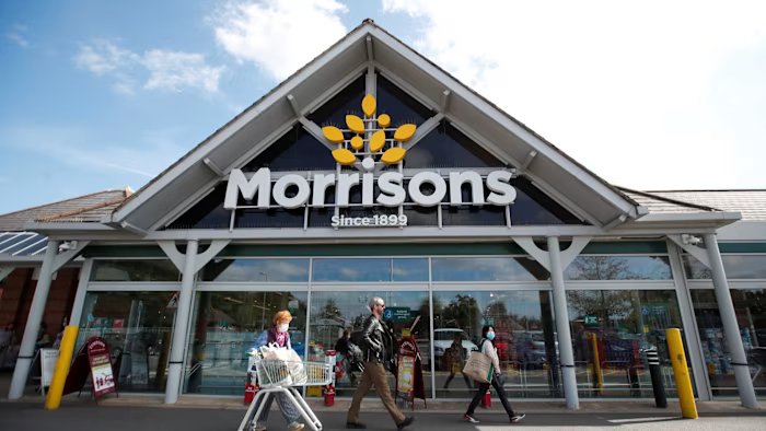 The entrance of a Morrisons supermarket in St Albans with people walking past and one person pushing a shopping cart.