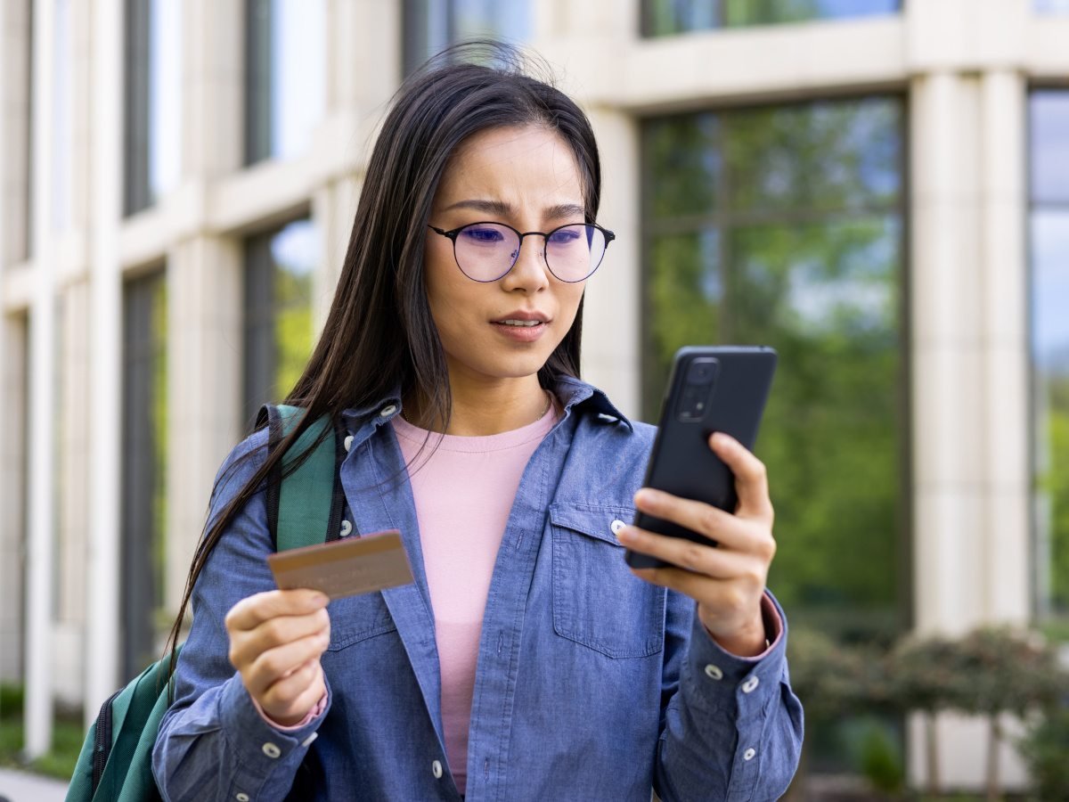 A woman with dark hair and glasses looking into her smartphone with an ATM card in the other hand.
