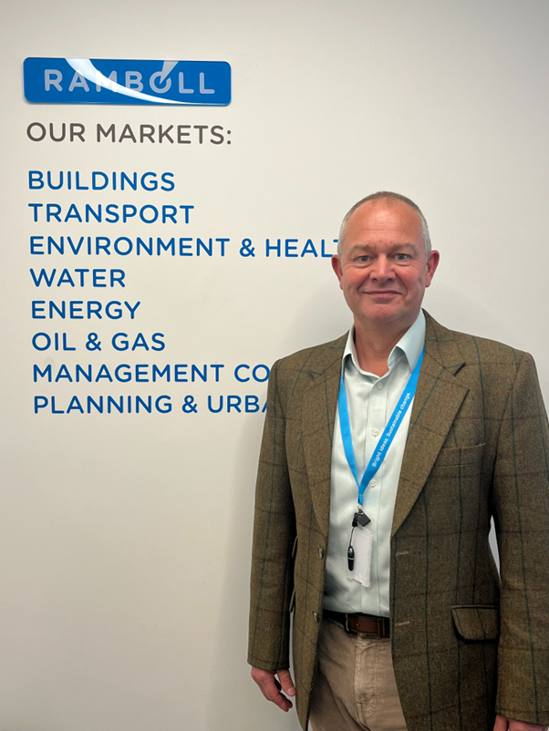 Portrait of Richard Graybrook standing beside a Ramboll wall display outlining its market sectors, marking his appointment to lead the company’s aviation market in the UK, Ireland, and Europe. He wears a blazer and lanyard, positioned against a white backdrop featuring Ramboll branding and listed sectors including transport, energy, and planning.
