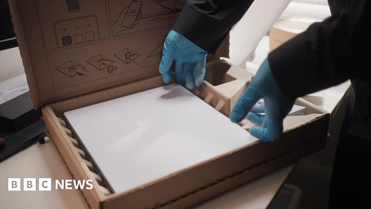 A white rectangular Starlink device rests inside a brown cardboard box. The hands of a person wearing thin blue plastic gloves and dark clothing are touching it at the edges, as if lifting it out of the box.