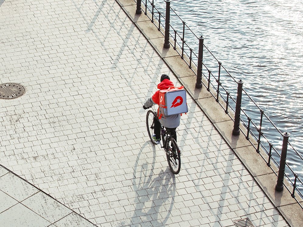 A person seen from afar wearing Delivery Hero uniform with a hot bag on a bicycle next to a body of water.