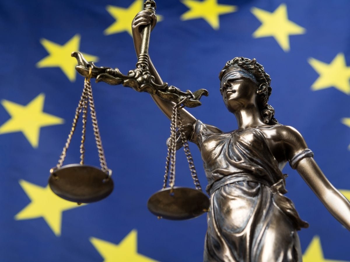 Bronze Lady Justice statue holding scales in front of a blurred European Union flag with yellow stars on a blue background.