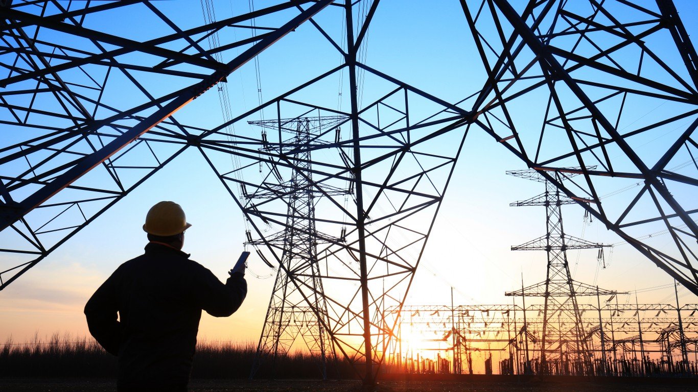 A silhouette of a worker in a yellow hard hat, viewed from behind, stands overlooking numerous high-voltage power transmission towers and lines against a vibrant sunset sky. The sky transitions from orange at the horizon to blue overhead, with additional substation infrastructure visible in the distance.